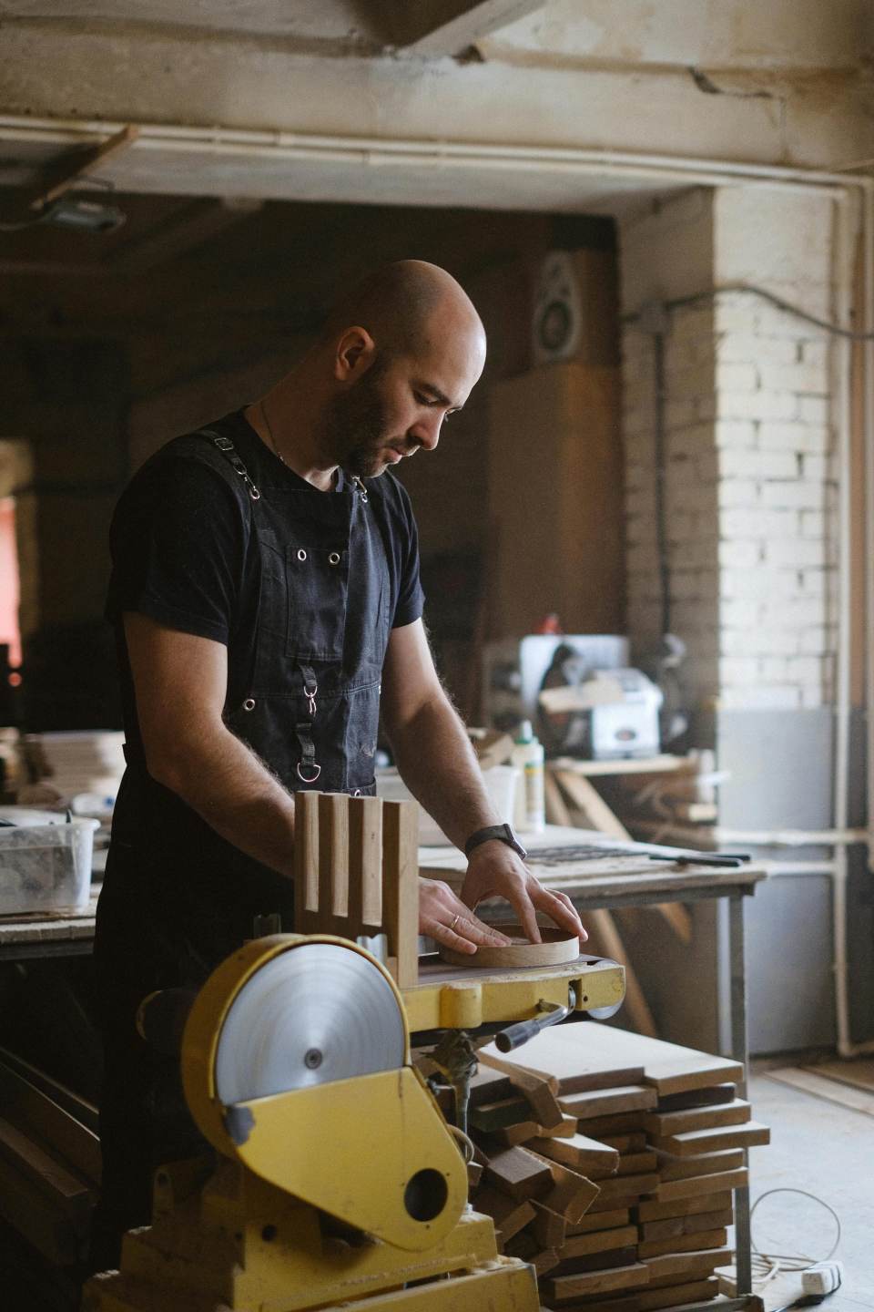 Vista lateral de un carpintero experto puliendo una pieza redonda de madera mientras trabaja con una lijadora de banda y disco en un taller con una pila de madera